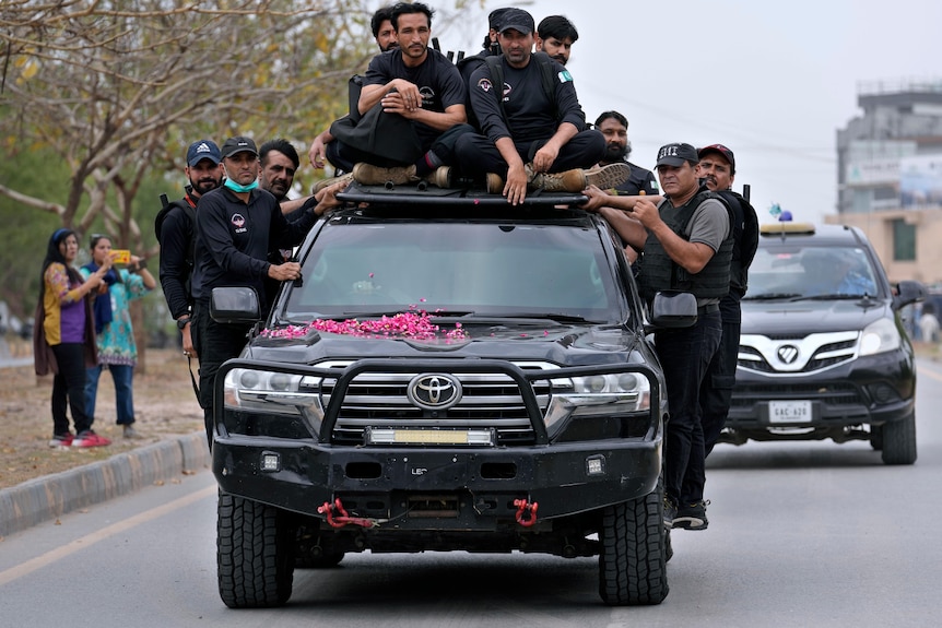 Security personnel in black uniforms hang on the side and sit on the roof of a black 4WD as it drives along a paved road.