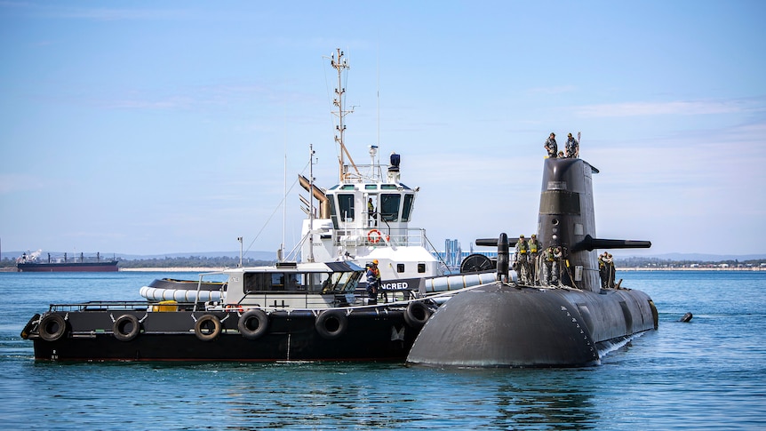 Submariners stand on a surfaced submarine, while two vessels are tethered to the boat