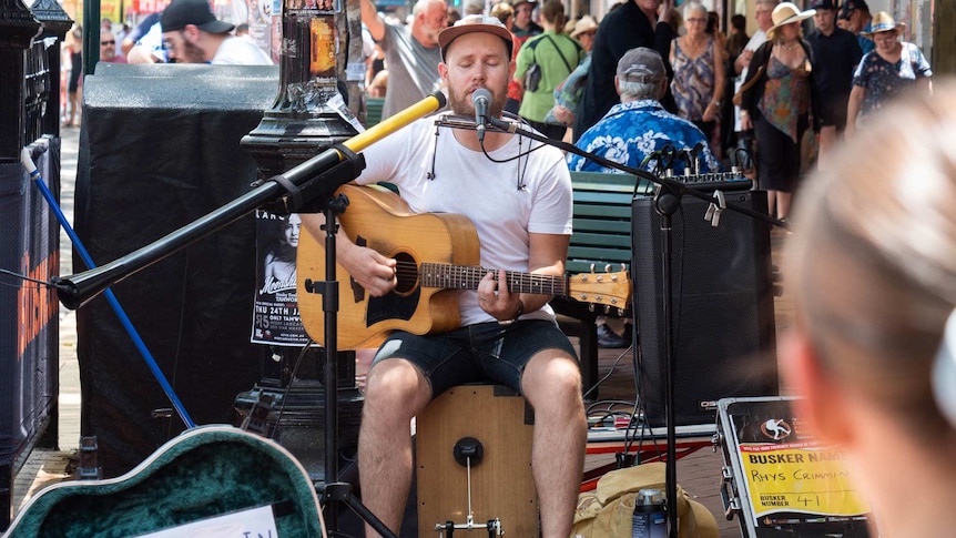 Rhys Crimmin plays guitar and sings into a microphone of Peel Street, guitar case in front of him