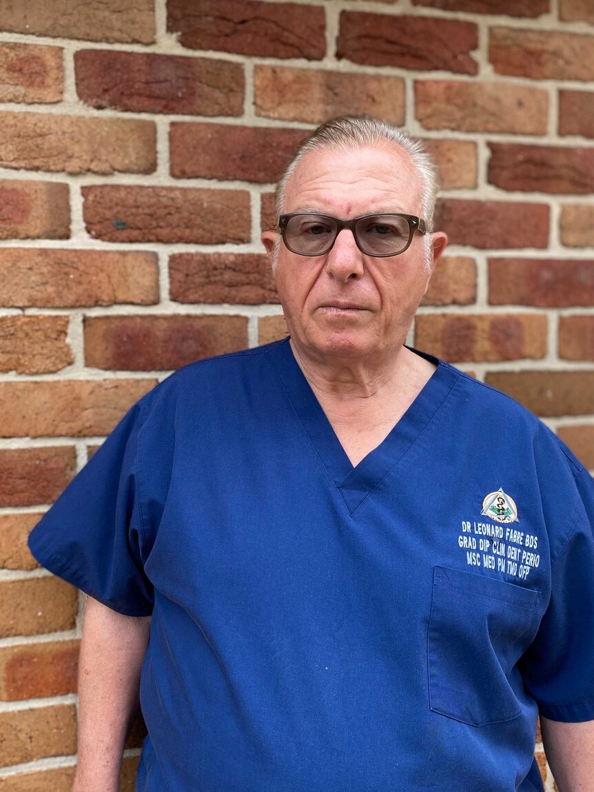 A man in a blue dentist uniform and tinted glasses stands against a brick wall