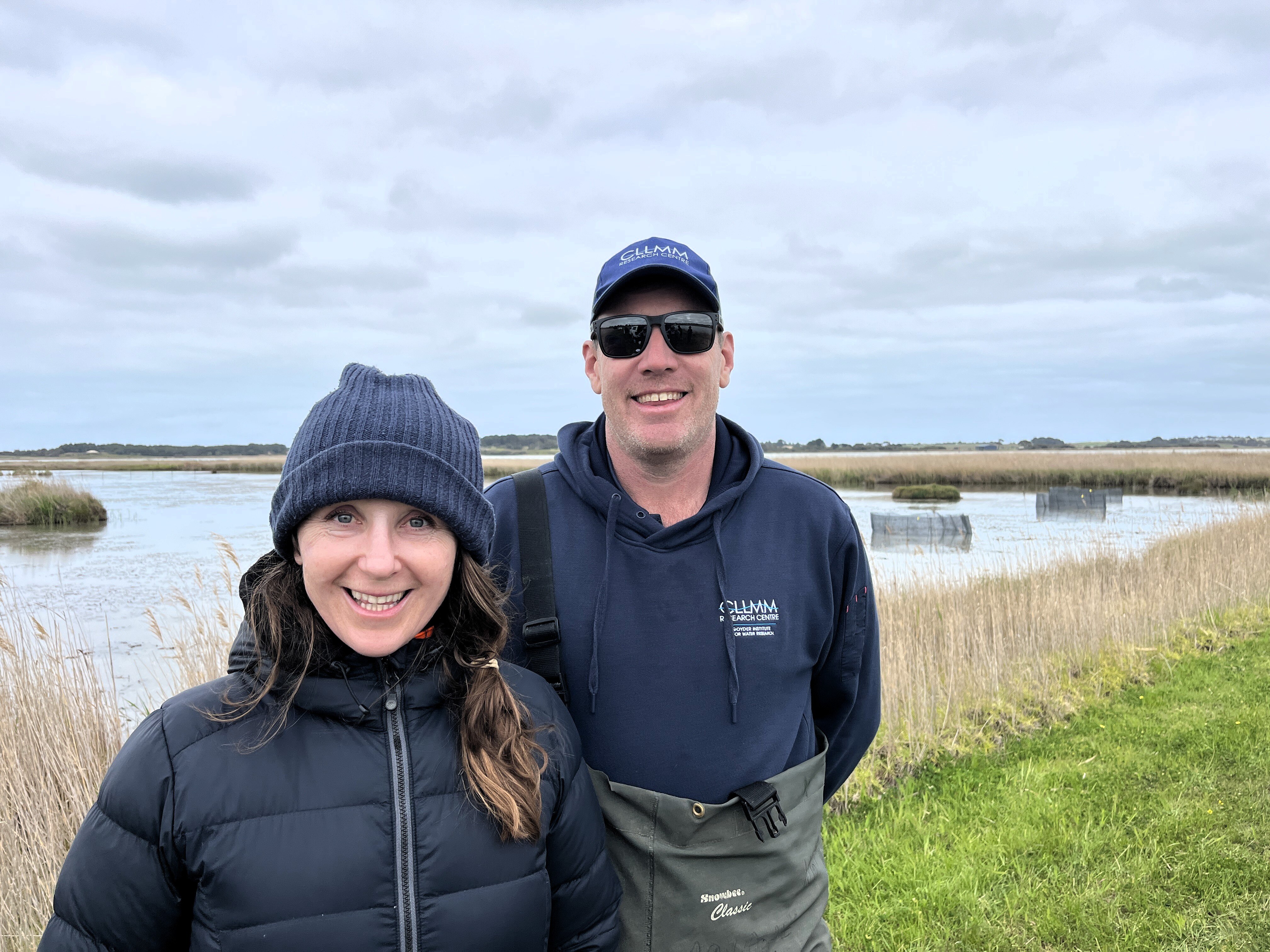 A man and a woman stand by a river with reeds and fish enclosures in the background.