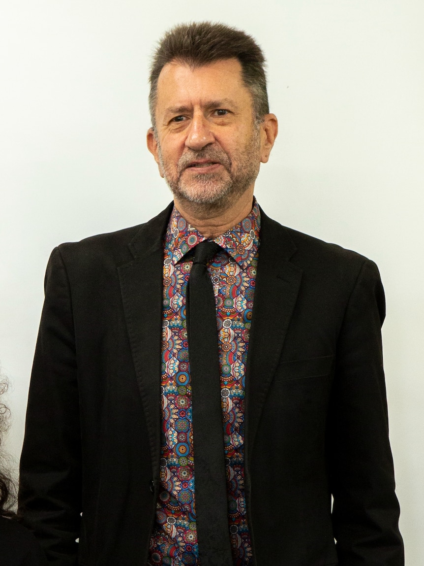 A closely cropped image of a man wearing a patterned shirt and black suit against a white backdrop.