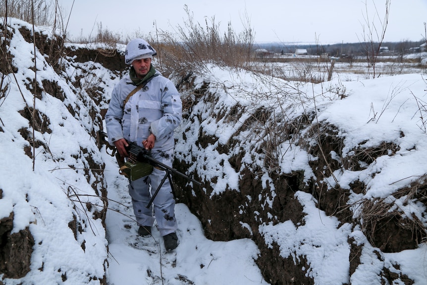 Man in military gear stands in a snowy trench, holding a gun. His camouflage is white 