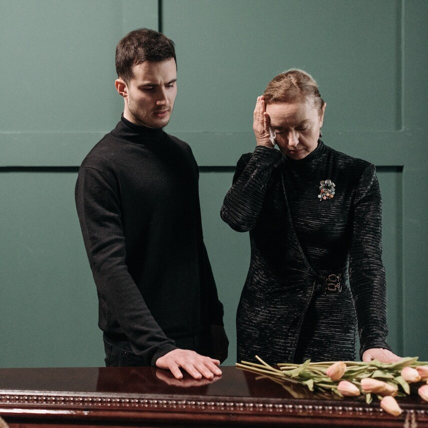 Adult son and his mother standing over a wooden coffin dressed in black