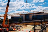 Wide shot of a construction site with orange cranes and fences, a glass building in the background and blue sky with clouds.