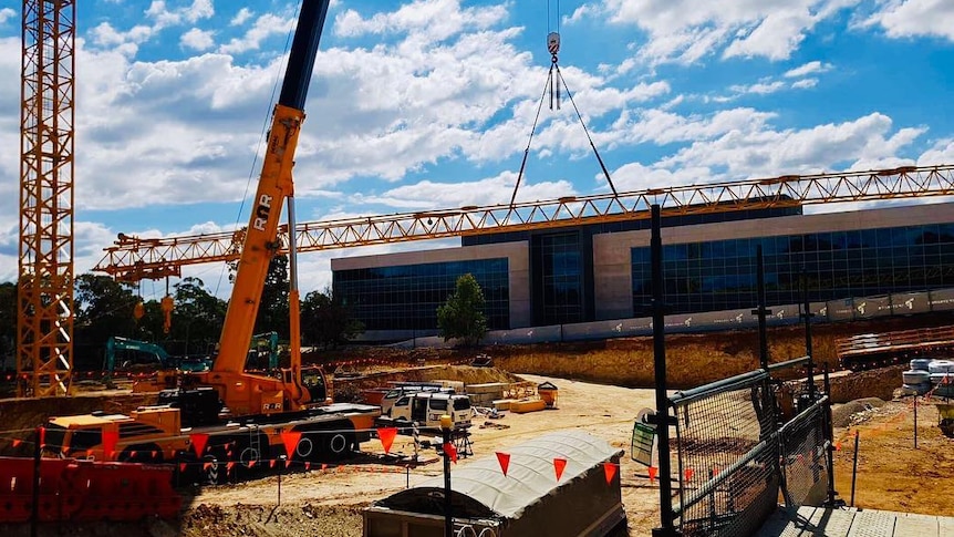 Wide shot of a construction site with orange cranes and fences, a glass building in the background and blue sky with clouds.