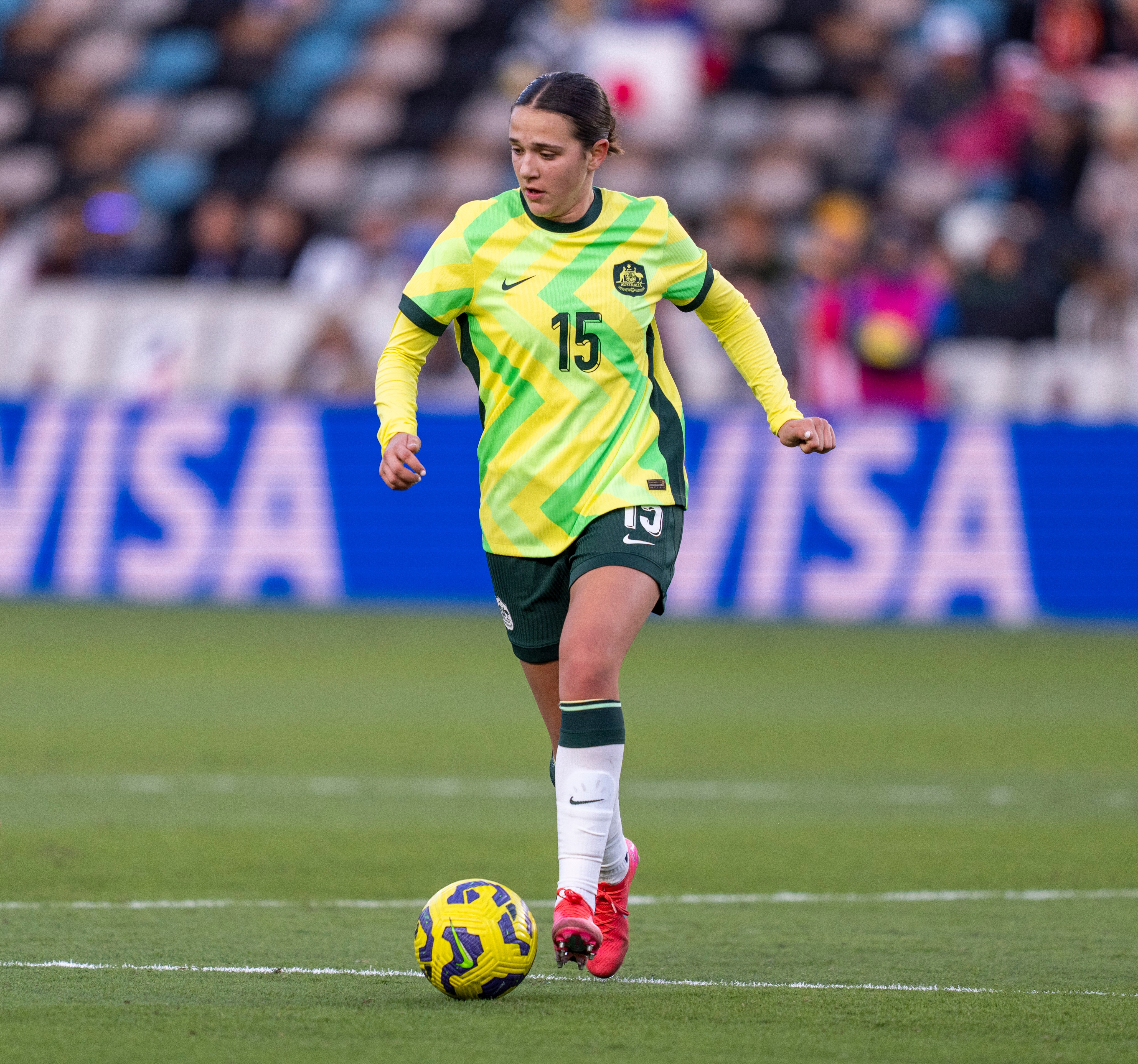 A woman with tied back brown hair in a yellow and green football kit with looks down with ball at feet 