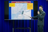 Man in military uniform speaks into microphone while hold pointer towards large screen showing map of Taiwan.