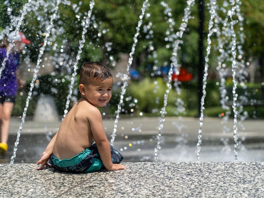 A toddler in green swimming shorts sits on the edge of a water fountain