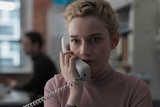 The actor Julia Garner in the film The Assistant, she's wearing a pink top, blonde hair, holding a landline phone up to her ear
