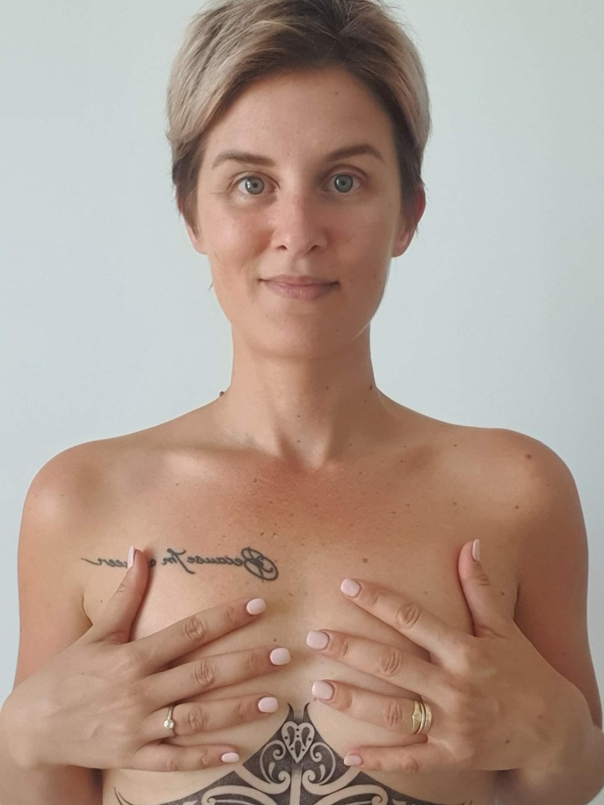 Myjanne stands topless with her hands over her breasts. She has a couple of tattoos, one on her chest, one between her breasts.