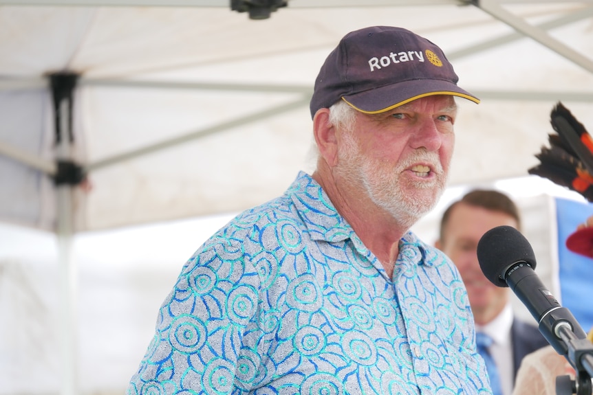 An older, bearded man in a cap speaks into a microphone.