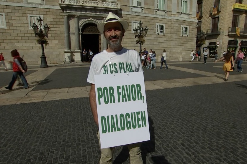 A man stands wearing a sandwich board saying "please talk" in Spanish.