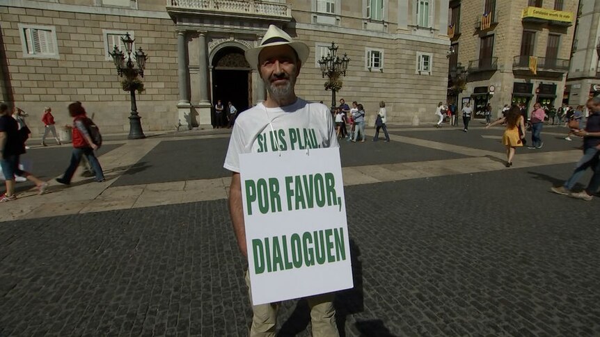 A man stands wearing a sandwich board saying "please talk" in Spanish.