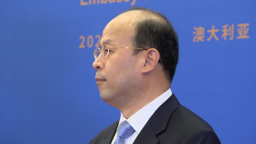 A profile shot of a middle-aged balding man wearing a suit and glasses, in front of a large backdrop.