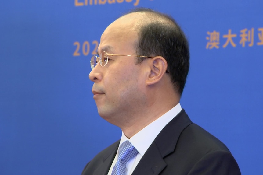 A profile shot of a middle-aged balding man wearing a suit and glasses, in front of a large backdrop.