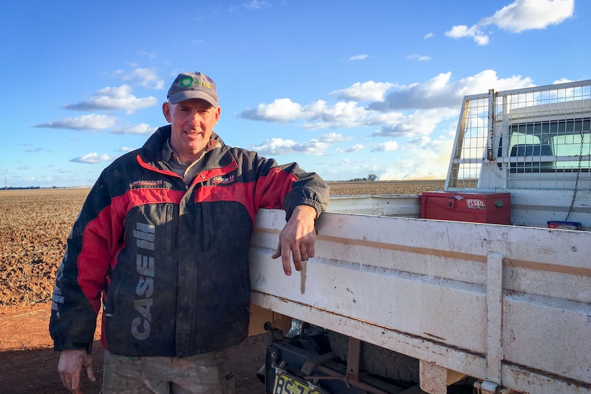 Man in blue hat leans on tray of his ute, next to a bare paddock.