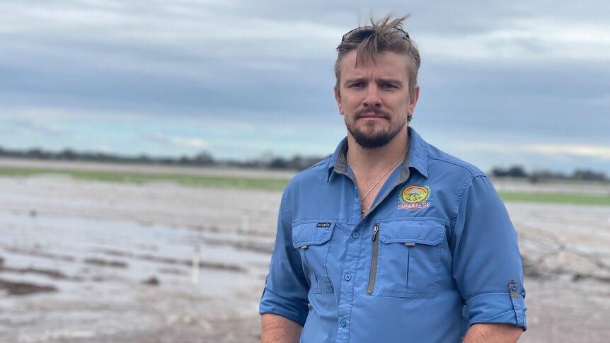 A fair-haired, neatly-bearded man in work gear stands in front of a flooded paddock, looking solemn.