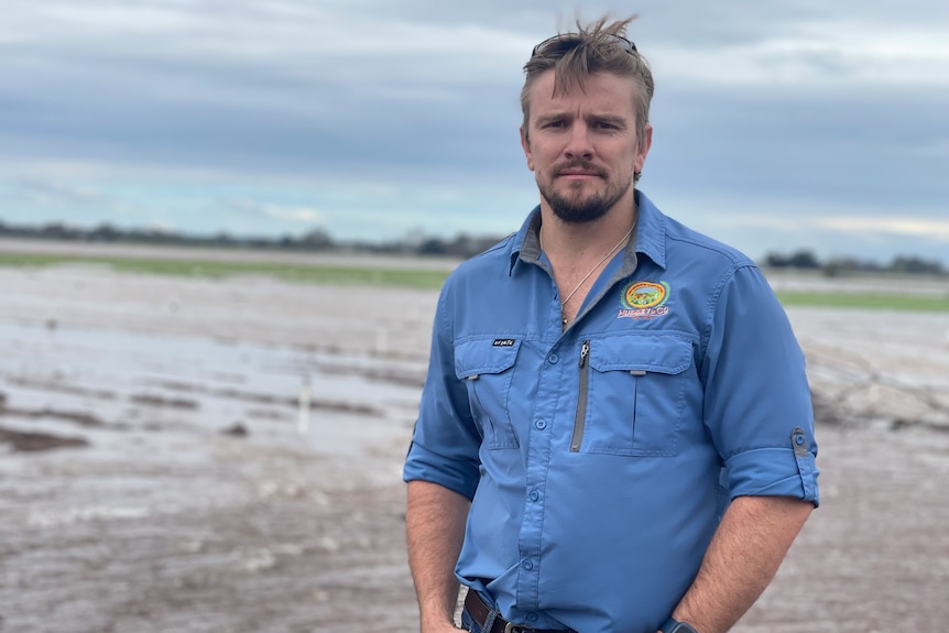 A fair-haired, neatly-bearded man in work gear stands in front of a flooded paddock, looking solemn.