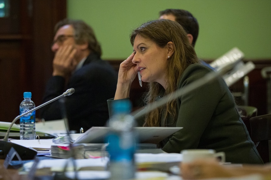 A woman with long brown hair and a black jacket sits at a desk with a microphone and paperwork. Her hand is resting on her face