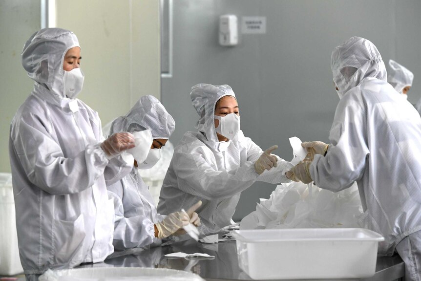 Workers in white hazmat suits and face masks hold an inspect partially constructed masks sitting on a silver bench.