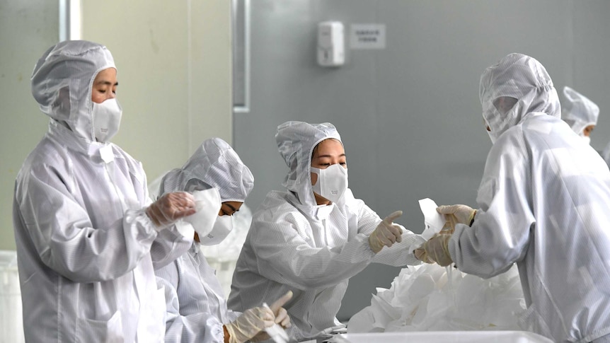 Workers in white hazmat suits and face masks hold an inspect partially constructed masks sitting on a silver bench.