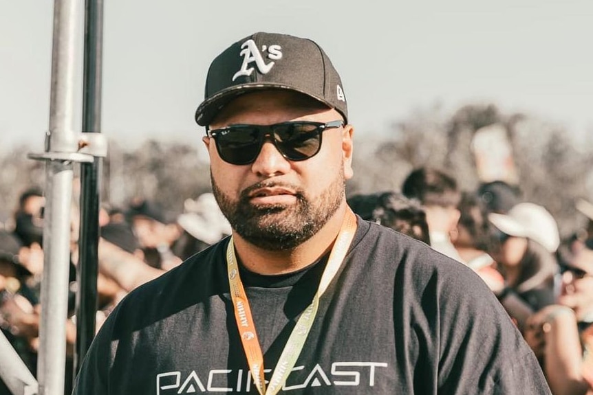 A tall pacific island man wearing a hat and sunglasses at a music festival with lanyard on