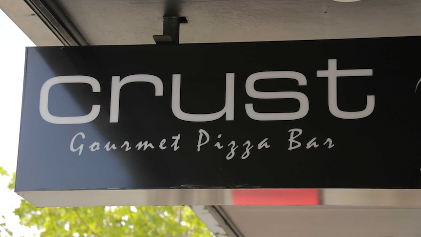 A black sign with the words "Crust Gourmet Pizza Bar" written in white hangs from an outdoor awning