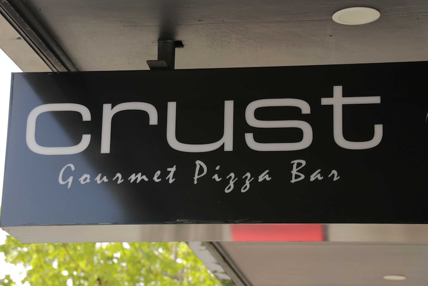 A black sign with the words "Crust Gourmet Pizza Bar" written in white hangs from an outdoor awning