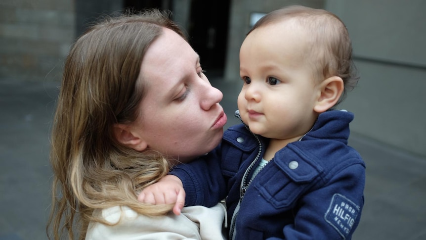 A close-up shot of a woman holding and kissing a young toddler in a blue jacket.