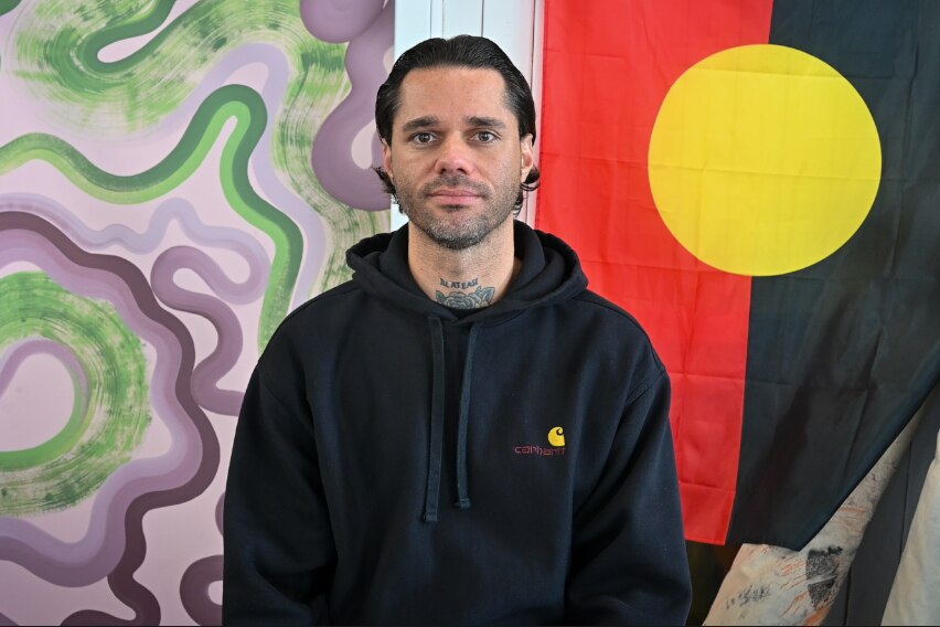 A man wearing a black hoodie sits in from of a purple and green artwork and the Aboriginal flag. 