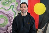 A man wearing a black hoodie sits in from of a purple and green artwork and the Aboriginal flag. 