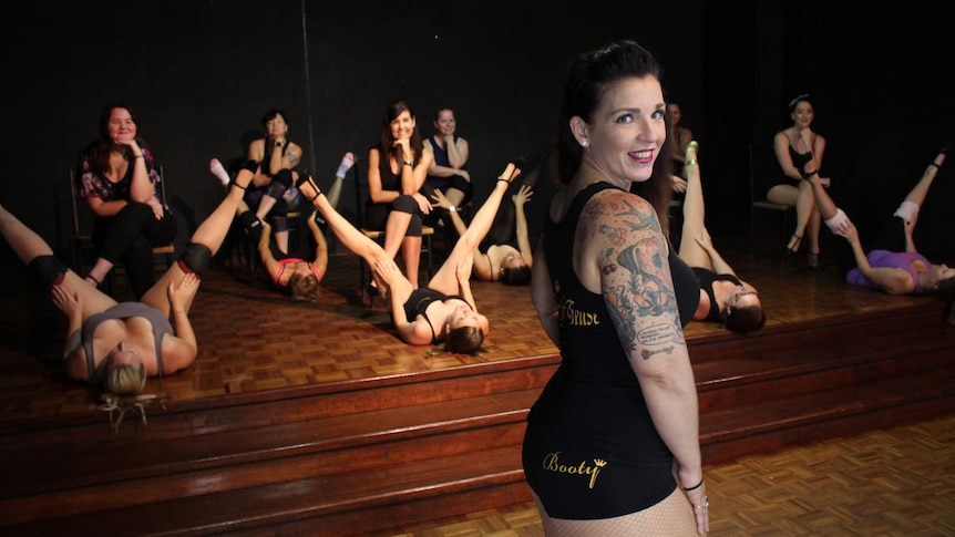 A woman with long hair and tattoos stands in front of a group of burlesque performers who are wearing training costumes