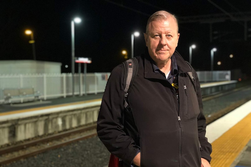 A man standing with black jacket, looking like he has come back from work, at night at the train station.