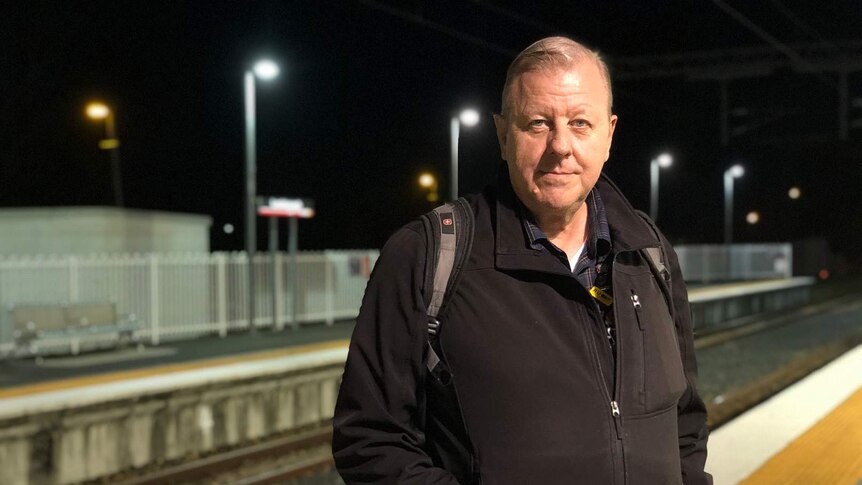 A man standing with black jacket, looking like he has come back from work, at night at the train station.