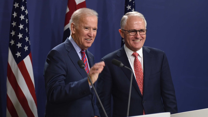 Joe Biden and Malcolm Turnbull stand smiling in front of US and Australian flags.