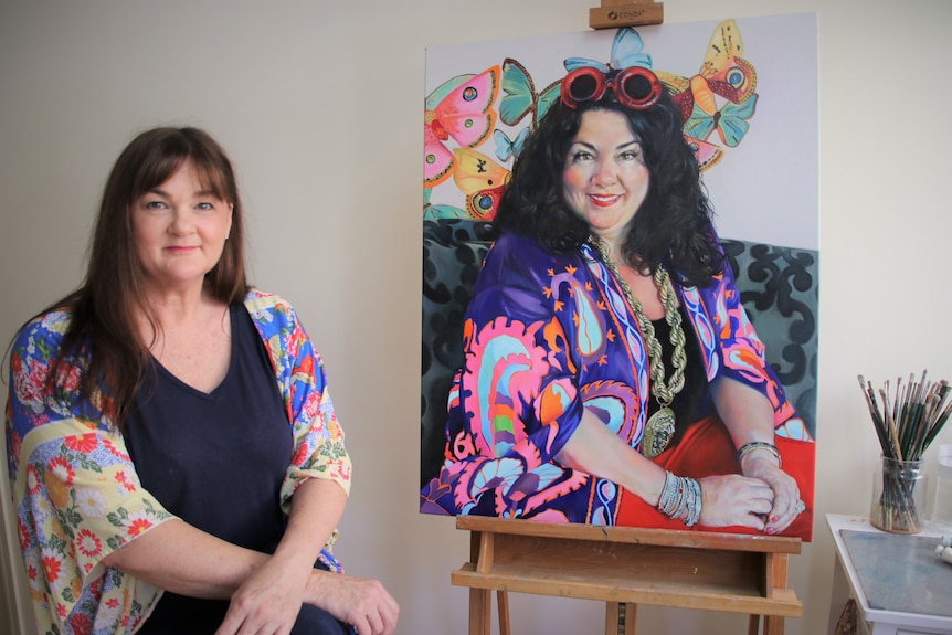 A woman sits next to a painted portrait.