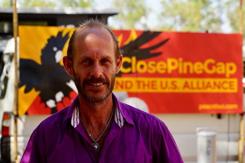 Paul Christie stands in front of a red and yellow 'Close Pine Gap' banner