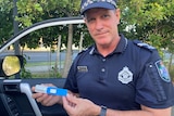 a male police officer looks at the camera while holding a small blue drug test kit 