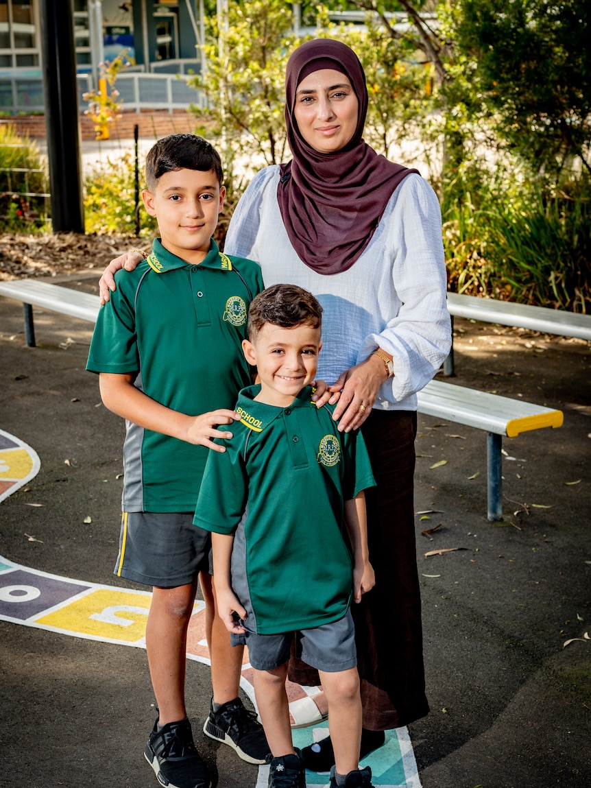 Ashty Fathi stands behind her two sons, smiling in the school grounds, in a portrait-style photo.