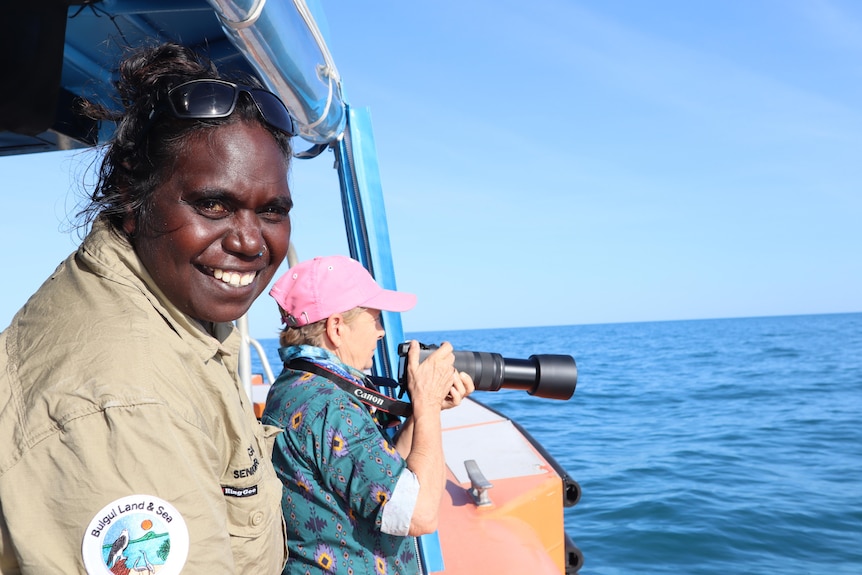 An Aboriginal woman in tan shirt smiles at the camera on the side of a boat, a woman in the background holds a camera.