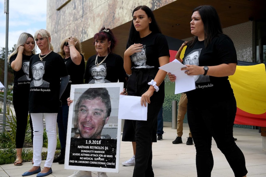 women in black shirts standing in front of the aboriginal flag holder a poster of a man