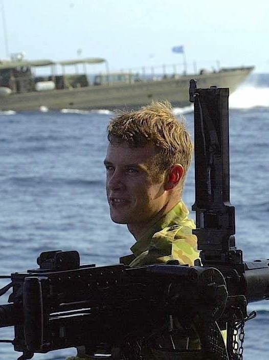 A man dressed in army camouflage uniform standing in behind a gun and standing in front of the ocean and a navy ship.