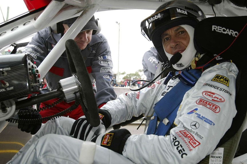 A man in a race car wearing a helmet and white overalls. A pit crew member is bending over in the open driver's door