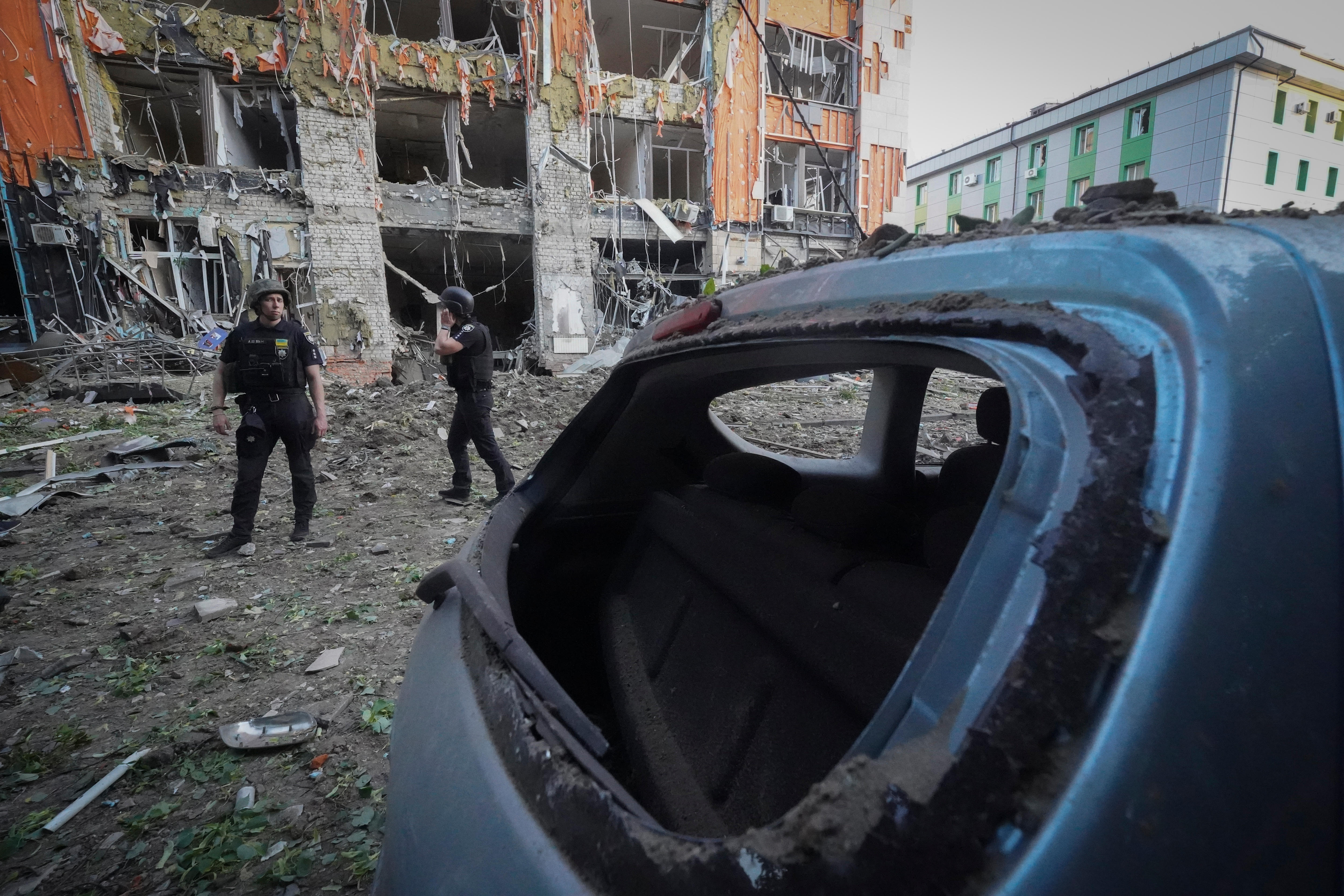 A bombed-out car sits in the street surrounded by debris from a nearby building, that stands damaged following a strike