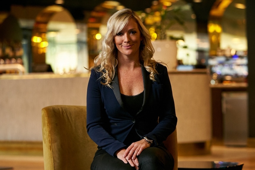 A blonde middle aged woman sitting down in a venue wearing a navy blazer and black shirt