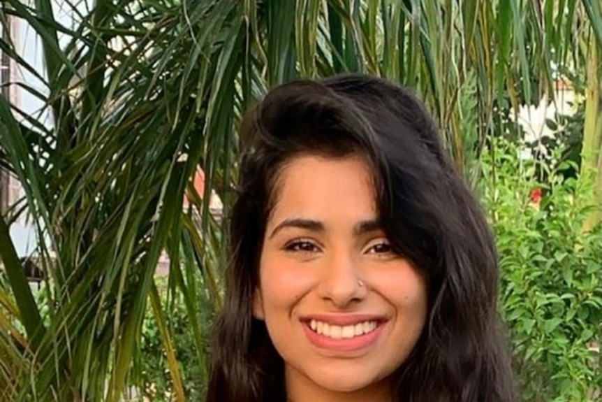 A young woman with long dark hair smiles while standing in front of a lush green garden