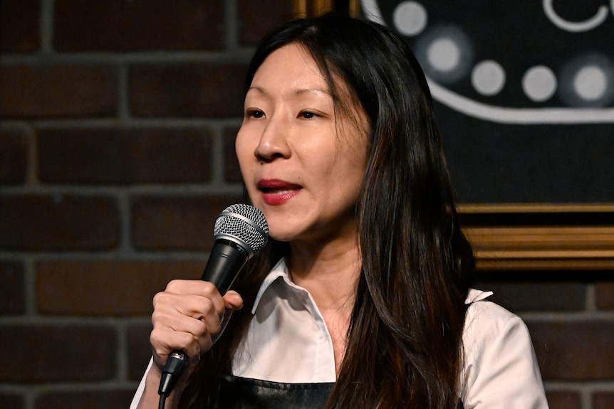 A woman of Asian heritage holding a microphone and speaking on stage. 