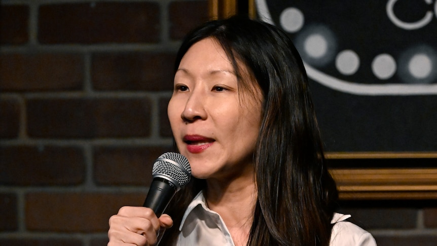 A woman of Asian heritage holding a microphone and speaking on stage. 