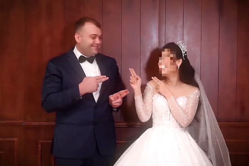 A man in a suit and bow tie and a woman in a wedding dress with her face blurred make gun signals at each other with their hands